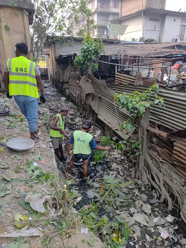 DMC sanitation workers in high-visibility vests clearing a drain between houses