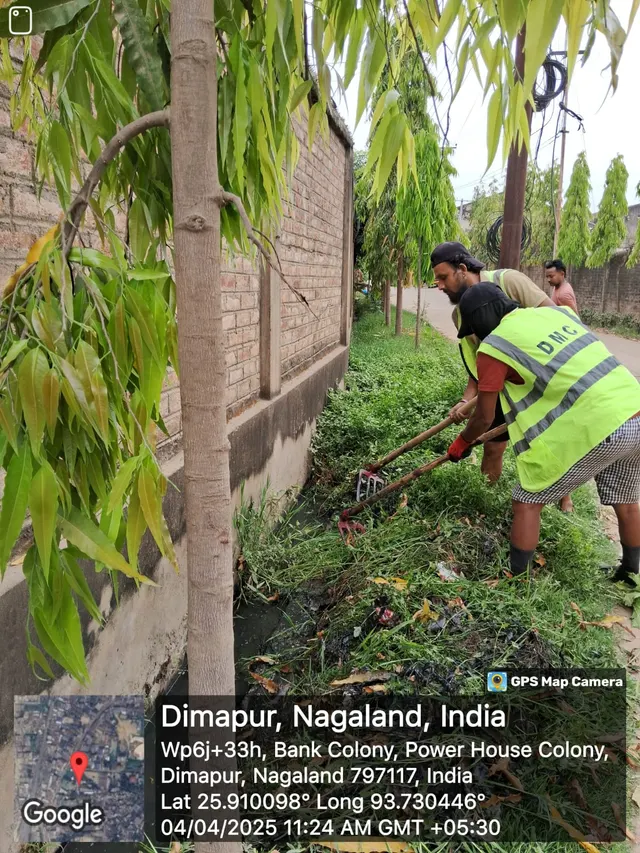 Sanitation workers clearing weeds from a drain along a wall